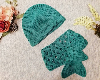 Puede incluir: Un gorro de punto de color azul verdoso y una cola de sirena de punto del mismo color. El gorro tiene un diseño de flor en un lado.