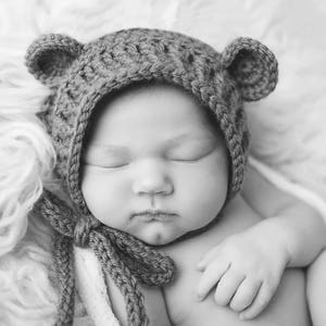 Puede incluir: Un beb&eacute; reci&eacute;n nacido con un gorro de oso de crochet marr&oacute;n con orejas ca&iacute;das, durmiendo sobre una manta blanca y esponjosa.