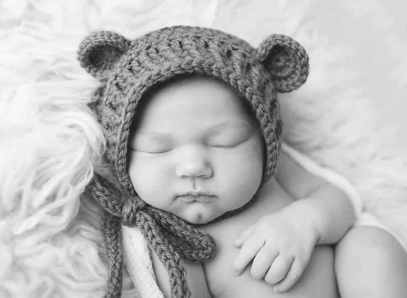 Puede incluir: Un beb&eacute; reci&eacute;n nacido con un gorro de oso de crochet marr&oacute;n con orejas ca&iacute;das, durmiendo sobre una manta blanca y esponjosa.