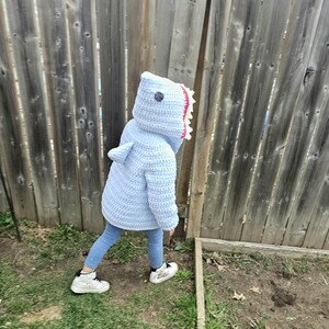 Puede incluir: Un ni&ntilde;o lleva un disfraz de tibur&oacute;n de crochet azul con una boca blanca y roja. El ni&ntilde;o est&aacute; de pie sobre la hierba frente a una valla de madera.