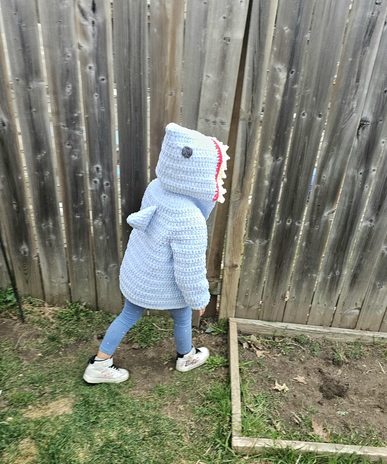 Puede incluir: Un ni&ntilde;o lleva un disfraz de tibur&oacute;n de crochet azul con una boca blanca y roja. El ni&ntilde;o est&aacute; de pie sobre la hierba frente a una valla de madera.