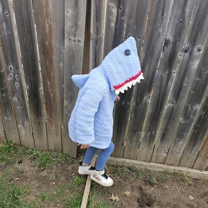 Puede incluir: Un disfraz de tibur&oacute;n de crochet azul claro con una boca blanca y roja. El disfraz tiene una capucha con una aleta de tibur&oacute;n en la espalda. Un ni&ntilde;o lleva el disfraz y est&aacute; de pie sobre una tabla de madera.