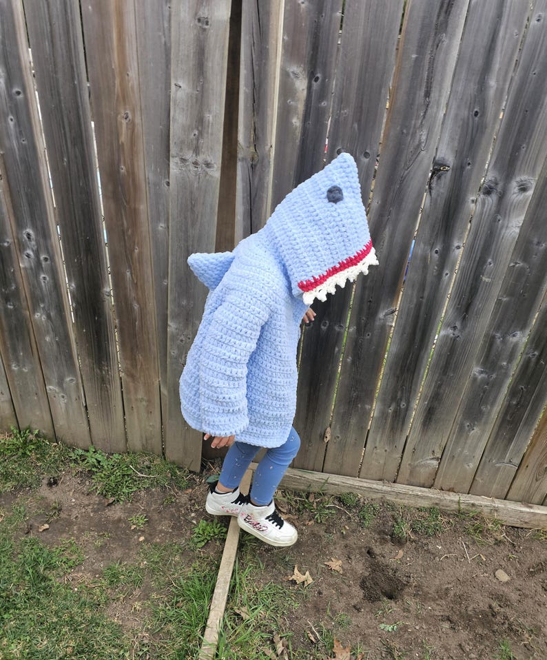 Puede incluir: Un disfraz de tibur&oacute;n de crochet azul claro con una boca blanca y roja. El disfraz tiene una capucha con una aleta de tibur&oacute;n en la espalda. Un ni&ntilde;o lleva el disfraz y est&aacute; de pie sobre una tabla de madera.