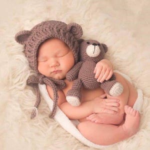 Puede incluir: Un beb&eacute; reci&eacute;n nacido con un gorro de oso de crochet marr&oacute;n con orejas, durmiendo en una manta blanca y esponjosa y sosteniendo un oso de peluche de crochet marr&oacute;n.
