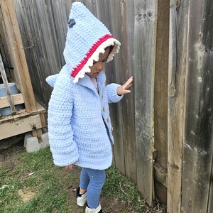 Puede incluir: Un ni&ntilde;o o ni&ntilde;a lleva una chaqueta de crochet azul con capucha de tibur&oacute;n con detalles de dientes rojos y blancos. La chaqueta tiene cierre de bot&oacute;n y el ni&ntilde;o o ni&ntilde;a lleva pantalones vaqueros azules y zapatillas blancas.