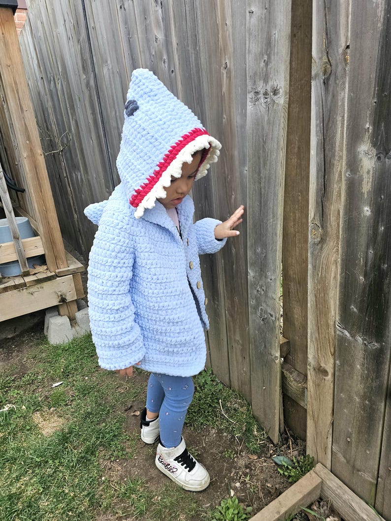 Puede incluir: Un ni&ntilde;o o ni&ntilde;a lleva una chaqueta de crochet azul con capucha de tibur&oacute;n con detalles de dientes rojos y blancos. La chaqueta tiene cierre de bot&oacute;n y el ni&ntilde;o o ni&ntilde;a lleva pantalones vaqueros azules y zapatillas blancas.