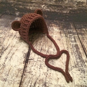 Puede incluir: Un gorro de beb&eacute; de ganchillo marr&oacute;n con orejas de oso y una larga corbata. El gorro est&aacute; sobre una superficie de madera.