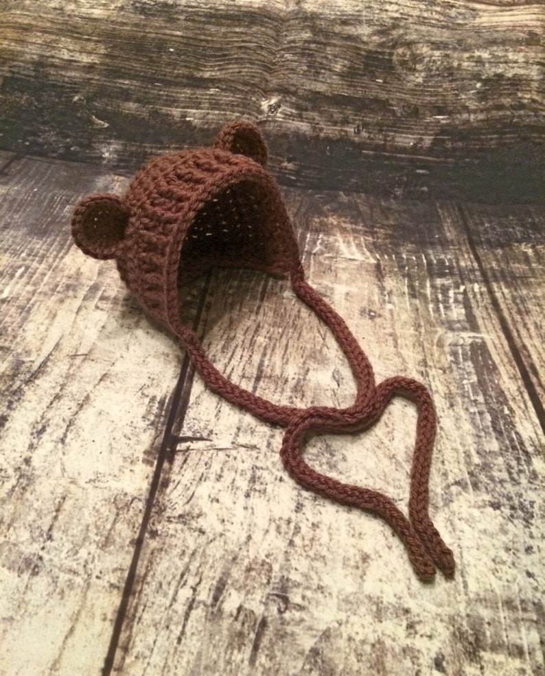 Puede incluir: Un gorro de beb&eacute; de ganchillo marr&oacute;n con orejas de oso y una larga corbata. El gorro est&aacute; sobre una superficie de madera.
