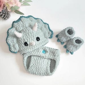 May include: A light blue crochet baby outfit, comprising a hat with horns, a diaper cover, and booties. The hat and booties are designed to resemble a triceratops dinosaur. The outfit features black eyes and a blue button.