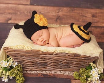 May include: A newborn baby wearing a black and yellow bee costume, sleeping in a wicker basket lined with a cream-colored blanket. The baby is wearing a black crocheted bee hat with a yellow flower on top and a matching yellow and black striped diaper cover.