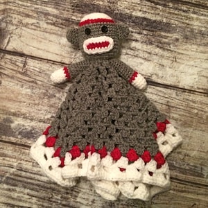 May include: A gray and white crocheted baby blanket with a red and white sock monkey attached. The blanket has a white crocheted border with red accents.