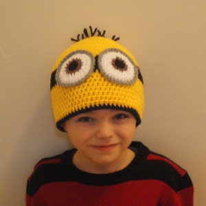May include: A child wearing a yellow crocheted Minion hat with black and white eyes. The hat has a black brim and is worn over a red and black striped shirt.
