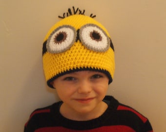 Puede incluir: Un niño con un gorro de Minion de crochet amarillo con ojos negros y blancos. El gorro tiene un borde negro y se lleva sobre una camisa a rayas rojas y negras.