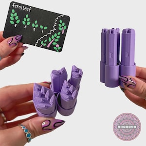 May include: A set of purple fern leaf-shaped craft tools. The tools are held by hands with painted nails. A small black card with the text "Fern/Leaf" and a diagram of fern leaves is also present.