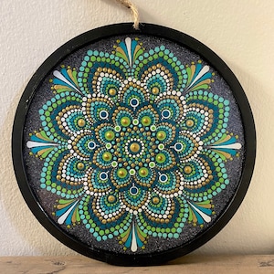 May include: A round wooden wall hanging with a mandala design painted in shades of green, blue, white, and gold. The mandala is made up of dots and circles.