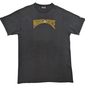 May include: Dark gray t-shirt with the text "VOLCOM STONE" in yellow arched lettering. The shirt has a crew neck and short sleeves. A small logo is visible on the upper back of the shirt.