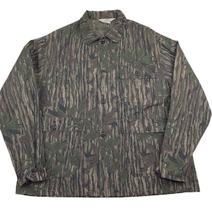 Puede incluir: Una camisa de camuflaje de manga larga con botones, dos bolsillos en el pecho y cuello. La camisa presenta un patrón de líneas verticales en tonos marrón, verde y beige, con detalles en forma de hojas.