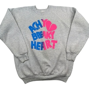 Puede incluir: Sudadera gris con un estampado gráfico rosa y azul que dice "achy breaky heart" con un gráfico de corazón roto.
