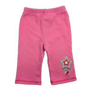May include: Pink baby pants with a star and monkey applique on the left leg. The pants have a red stitched hem and waistband. The star applique is green, blue, and red, with a yellow star in the center. The monkey is red and brown.