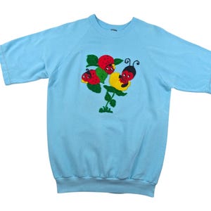 Op de afbeelding: Een lichtblauw sweatshirt met korte mouwen met een geborduurd ontwerp van drie lieveheersbeestjes op een groene plant met rode aardbeien.