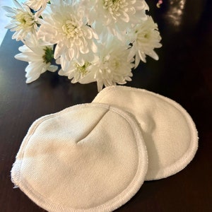 May include: Two white cotton reusable breast pads with a stitched edge. The pads are on a dark brown surface.