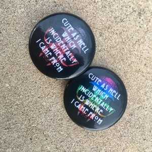 May include: Two black circular badges with the text "CUTE AS HELL WHICH INCIDENTALLY IS WHERE I CAME FROM" in white, red, and rainbow hues. They rest on a textured, light brown surface.