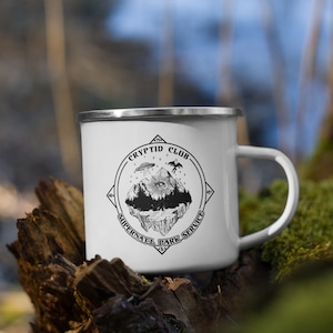 Peut inclure: Mug en émail blanc avec un bord et une anse argentés. Le mug présente un graphisme noir et blanc d'une île flottante avec des arbres, un œil et un OVNI, avec le texte "CRYPTID CLUB SUPERNATURAL PARK SERVICE" dans un design circulaire.