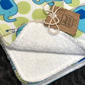 May include: A white, soft, and fluffy baby blanket with a blue and green elephant print. The blanket is folded in half and has a tag that says "Baby Hand Made USA".