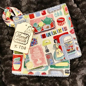 Puede incluir: Dos posavasos de tela hechos a mano con ribete azul y blanco. Los posavasos presentan un tema de cocina vintage colorido con imágenes de pasteles, un refrigerador y una tetera. Los posavasos están etiquetados como "Hand Made KIDS".