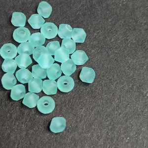 May include: A pile of light blue glass beads with a faceted design. The beads are scattered on a dark grey surface.
