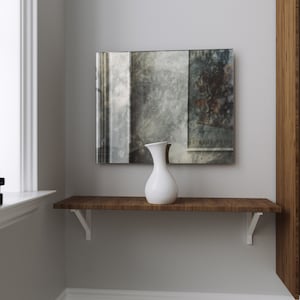 May include: A white ceramic pitcher sits on a wooden shelf in front of a wall with a framed abstract painting. The shelf is attached to a wall with a wooden bookcase.