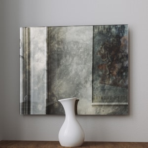 May include: A white ceramic vase with a rounded body and a narrow neck sits on a wooden surface in front of a large, rectangular mirror with a distressed, aged look. The mirror reflects a blurry image of a room with a window and a poster.
