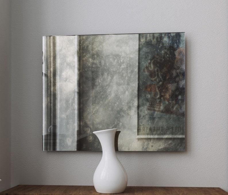 May include: A white ceramic vase with a rounded body and a narrow neck sits on a wooden surface in front of a large, rectangular mirror with a distressed, aged look. The mirror reflects a blurry image of a room with a window and a poster.