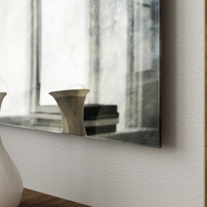 May include: A white ceramic pitcher with a rounded base and a wide mouth sits on a wooden surface in front of a large, rectangular mirror with a distressed, aged finish. The mirror reflects a blurry image of a vase and other objects.
