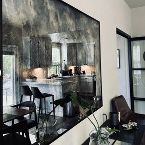 May include: A large, antique-style mirror with a black frame reflects a kitchen with white cabinets, a stainless steel sink, and a black countertop. The mirror is hanging on a white wall in a room with a black table and a brown leather chair.
