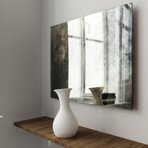 May include: A white ceramic vase with a rounded body and a narrow neck sits on a wooden shelf in front of a large, rectangular mirror with a distressed, aged finish. The mirror reflects a window with white blinds.