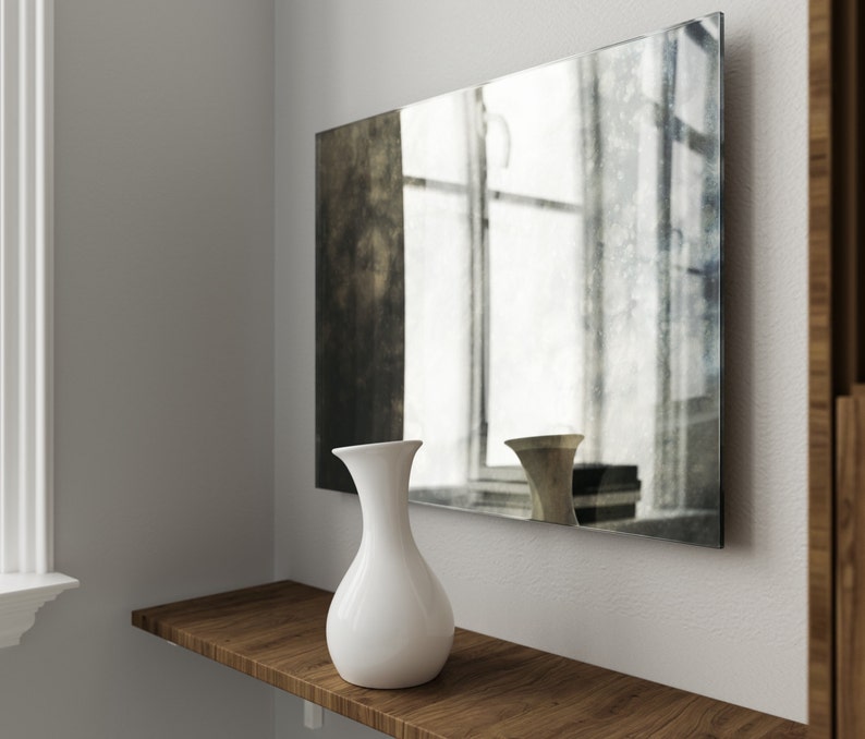 May include: A white ceramic vase with a rounded body and a narrow neck sits on a wooden shelf in front of a large, rectangular mirror with a distressed, aged finish. The mirror reflects a window with white blinds.