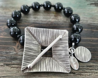 May include: A silver square bracelet with a textured design and a black onyx bead chain. The bracelet has a small silver charm with the text "Brenda Rae" on it.