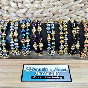 May include: Seven multi-colored beaded bracelets with gold accents. The bracelets are displayed on a wooden surface with a sign that reads "Brenda New Jewelry We don't do boring!"