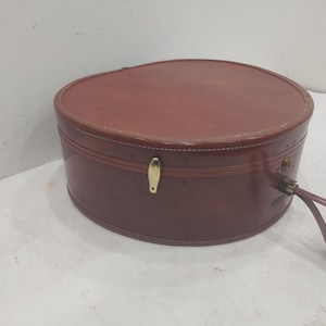 May include: A vintage, oval-shaped hat box in a rich, reddish-brown colour. The box is made of a leather-like material with a stitched seam around the top and bottom. It features a gold-coloured clasp and a matching handle.