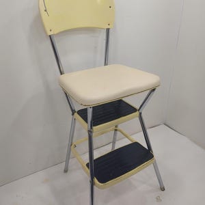 Puede incluir: Silla taburete vintage con asiento, respaldo y marco color crema. Los dos escalones son negros con superficie texturizada. Las patas son cromadas. Una pieza práctica y elegante para alcanzar lugares altos.