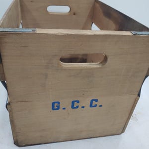 Op de afbeelding: Een vintage houten krat met uitgesneden openingen aan de zijkanten om te dragen. De krat is gemaakt van lichtgekleurd hout en verstevigd met metalen beugels. De letters "G.C.C." zijn blauw op de voorkant geschilderd.