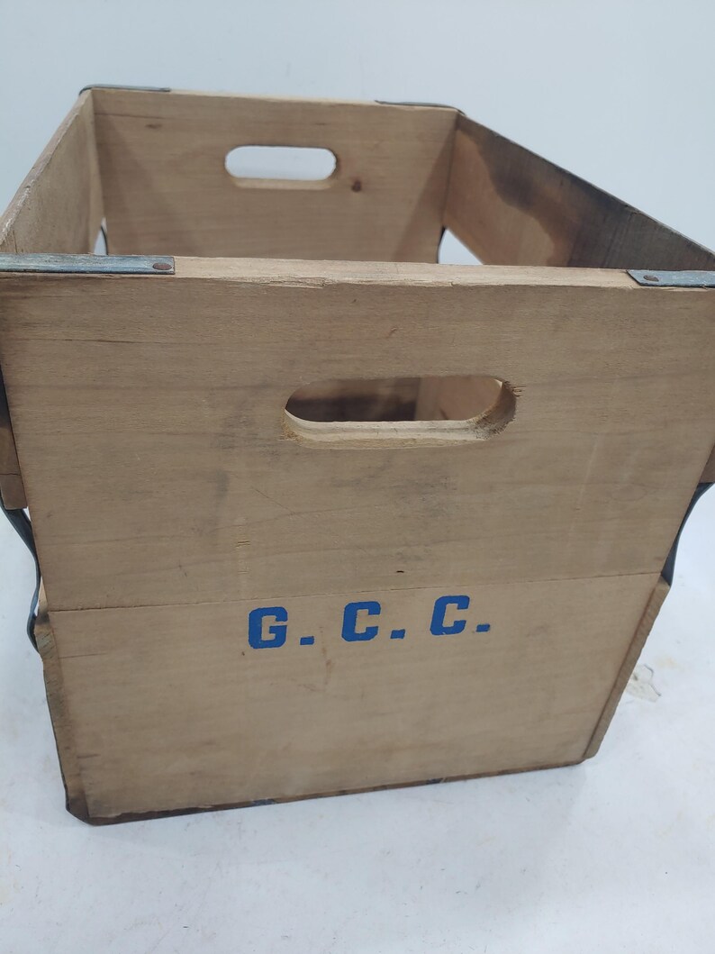 Op de afbeelding: Een vintage houten krat met uitgesneden openingen aan de zijkanten om te dragen. De krat is gemaakt van lichtgekleurd hout en verstevigd met metalen beugels. De letters "G.C.C." zijn blauw op de voorkant geschilderd.
