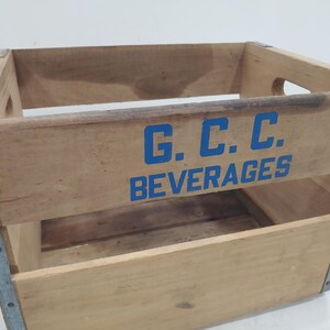 Op de afbeelding: Houten drankkrat met blauwe letters "G.C.C. BEVERAGES". De krat heeft metalen hoekverstevigingen en handgrepen aan de zijkanten. Het hout heeft een natuurlijke afwerking met gebruikssporen.