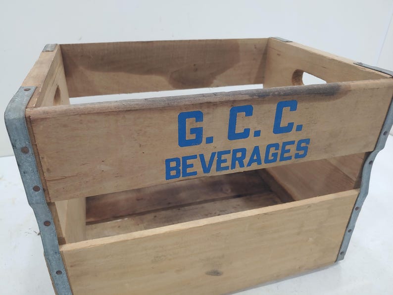 Op de afbeelding: Houten drankkrat met blauwe letters "G.C.C. BEVERAGES". De krat heeft metalen hoekverstevigingen en handgrepen aan de zijkanten. Het hout heeft een natuurlijke afwerking met gebruikssporen.