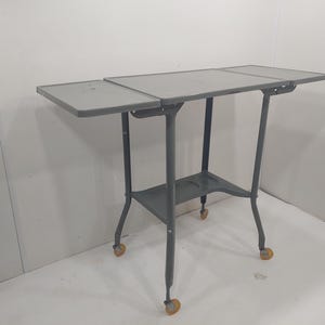 Może przedstawiać: Szary metalowy wózek do serwowania z prostokątnym blatem i dolną półką. Wózek ma cztery koła i jest podparty cienkimi, pochylonymi nogami. Blat ma dwa składane skrzydła, a wózek ma około 76 cm długości.