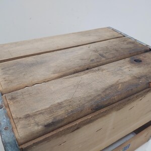 Op de afbeelding: Houten krat met metalen hoekverstevigingen. De krat is gemaakt van lichtbruin hout met zichtbare nerf en donkere vlekken. De bovenkant van de krat bestaat uit drie houten planken.