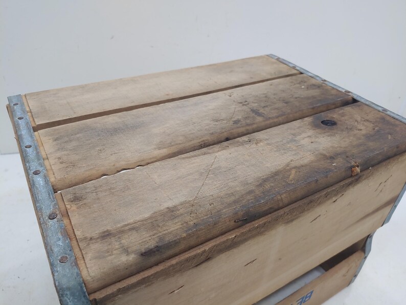 Op de afbeelding: Houten krat met metalen hoekverstevigingen. De krat is gemaakt van lichtbruin hout met zichtbare nerf en donkere vlekken. De bovenkant van de krat bestaat uit drie houten planken.