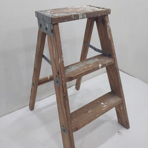 May include: A two-step wooden step stool with a top platform. The wood has a natural finish with visible grain and paint splatters. Metal brackets and hardware secure the frame. The stool is approximately 60 cm tall.