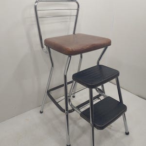Puede incluir: Un taburete con escalón cromado vintage con asiento y respaldo marrón. El taburete tiene un escalón negro y un marco cromado. El diseño es una combinación de taburete y escalera, adecuado para alcanzar lugares altos.